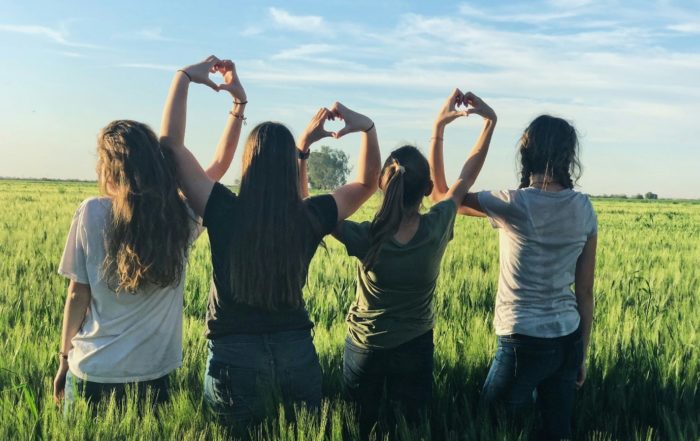 Four young women in a field making love heart symbols with their hands, for “Roses in the Snow: Supporting Young Women’s Wellbeing’’